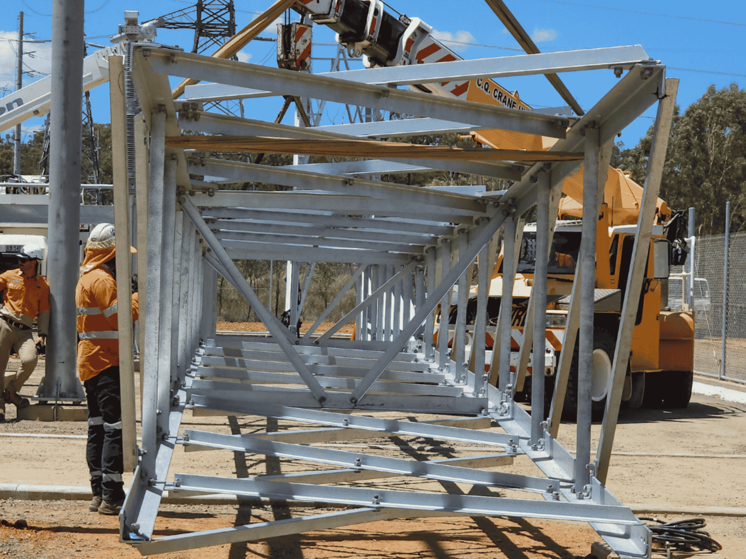 A galvanised steel support structure is lifted into position by crane at a power substation site, with workers nearby.